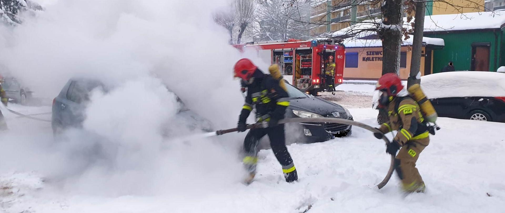 Palący się samochód gaszą strażacy ubrani w sprzęt ochrony układu oddechowego. W tle widać samochód pożarniczy i strażaka sterującego pracą autopompy. Obok pojazdu stoi inny samochód osobowy.