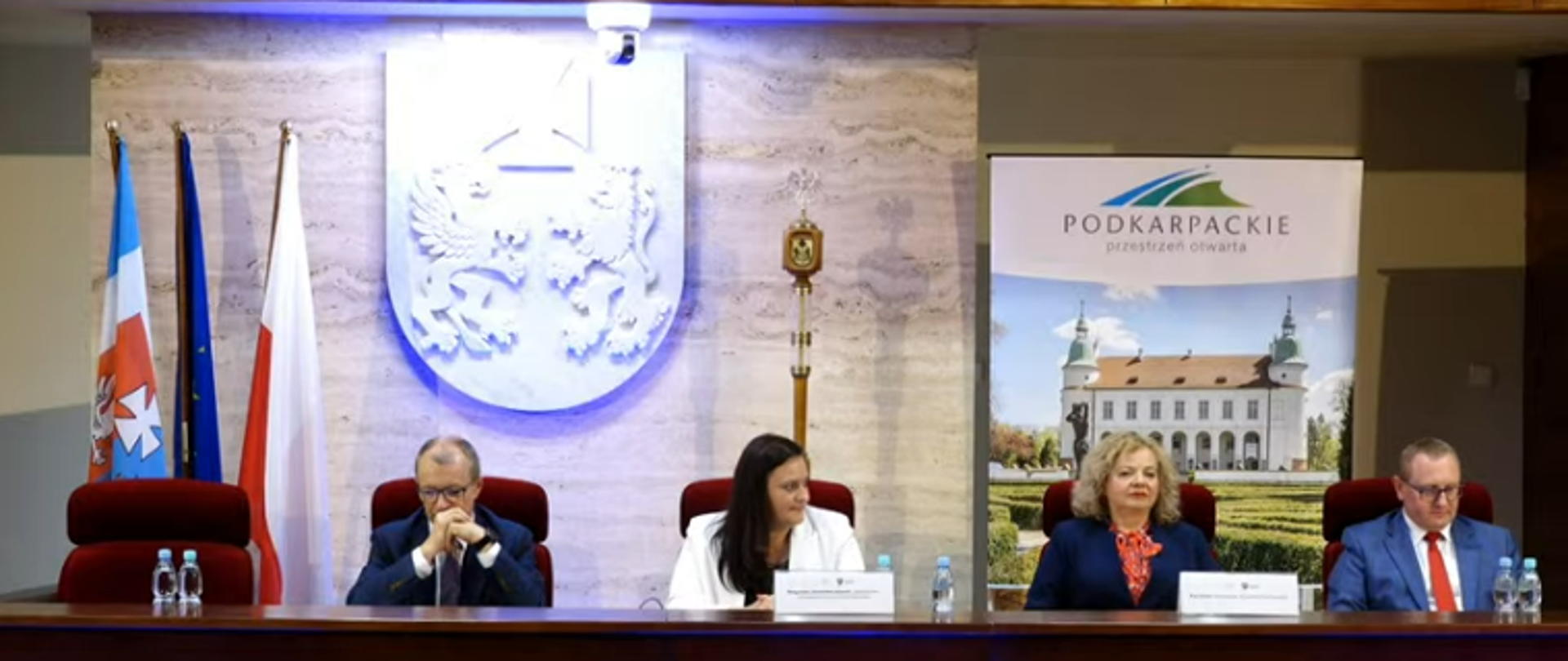 Za stołem konferencyjnym siedzą 4 osoby, w środku dwie kobiety, po bokach mężczyźni. Za nimi godło woj. podkarpackiego, banner ze zdjęciem pałacu i podpisem Podkarpacie oraz flagi Polski i UE
