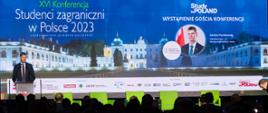 Widok z tyłu na zaciemnioną salę, na ustawionych w rzędy krzesłach siedzą ludzie, przed nimi na oświetlonym podwyższeniu przy mównicy stoi wiceminister Piontkowski i mówi do mikrofonu, za nim wielki ekran z napisem XVI Konferencja „Studenci zagraniczni w Polsce 2023” i Study in Poland - wystąpienie gościa konferencji.