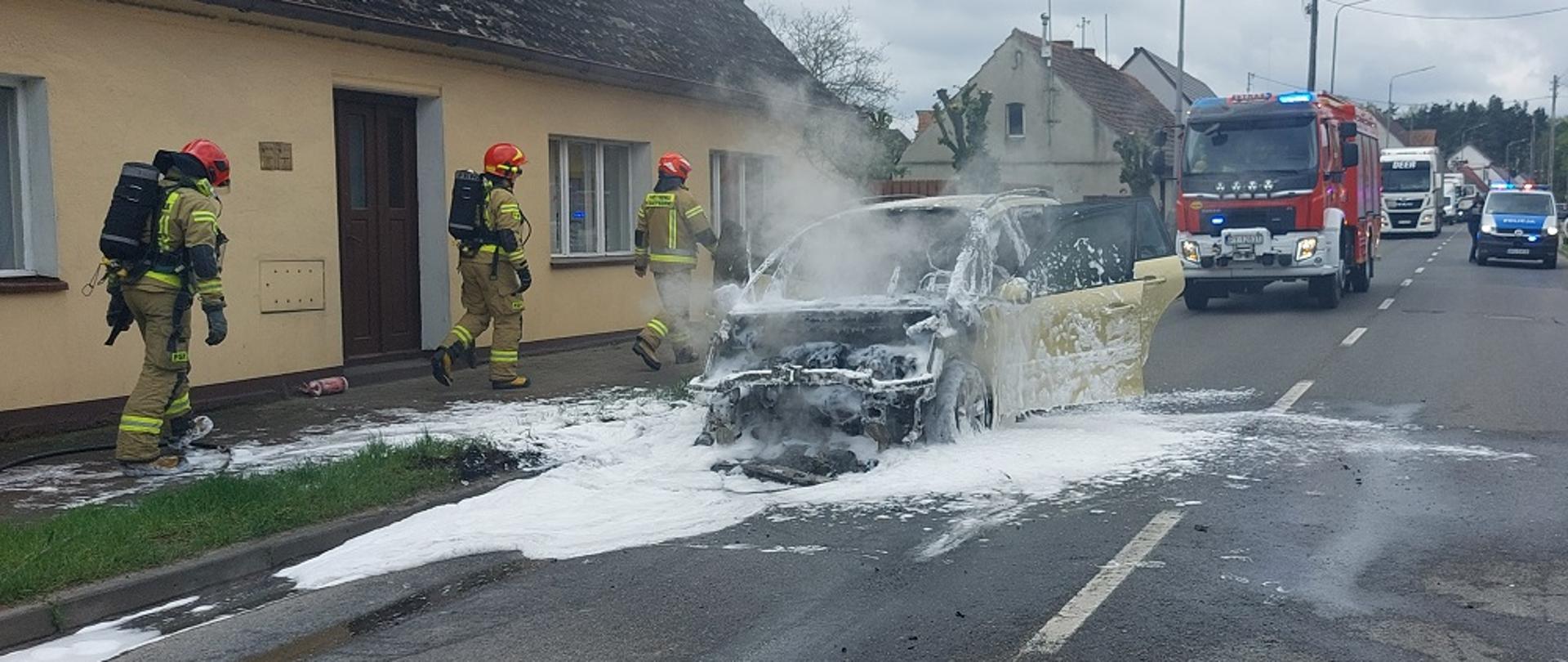 Zdjęcie przedstawia strażaków gaszących pożar samochodu osobowego stającego na środku drogi w miejscowości Margonin.
W tle jezdnia i budynki.
