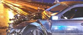 Na zdjęciu widać uszkodzony pojazd BMW. Uszkodzony przód pojazdu. Pogoda deszczowa. Pora nocna. W tyle niebieskie barierki drogowe. Oświetlenie uliczne w kolorze pomarańczowym.