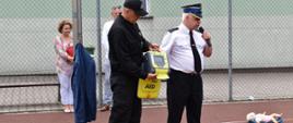 Boisko Zespołu Szkolno- Przedszkolnego w Zielonej Wsi. Dwaj strażacy prezentują automatyczny defibrylator zewnętrzny AED. Po prawej stronie leży fantom "niemowlak"