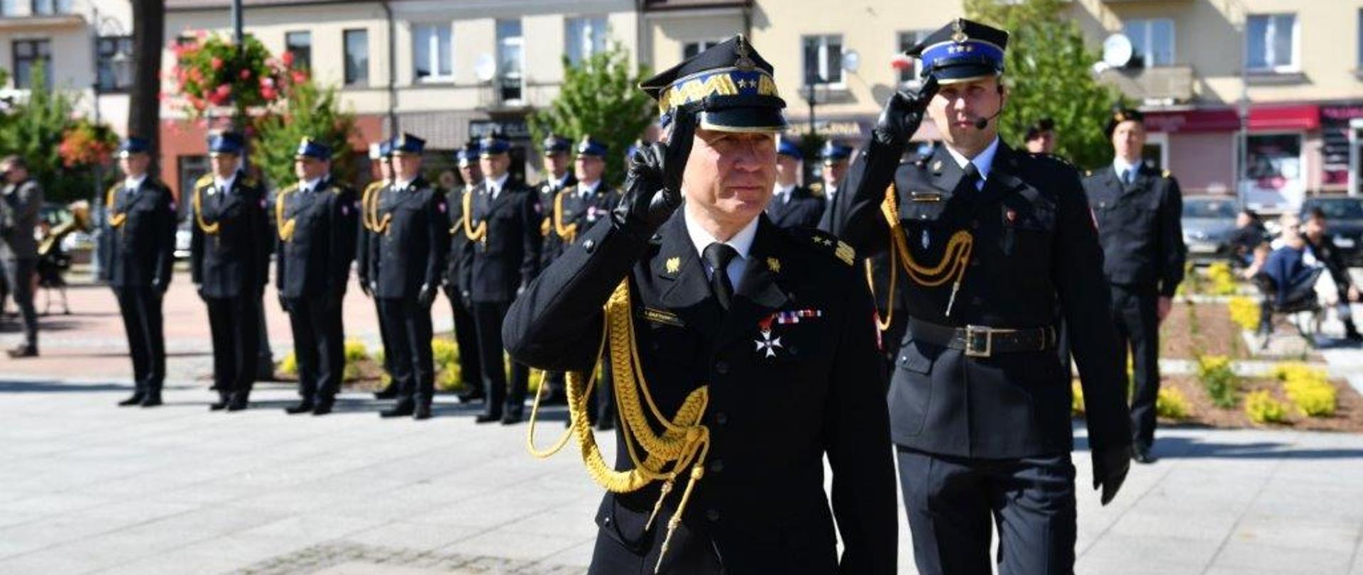 Komendant główny PSP a za nim dowódca uroczystości w mundurach galowych maszerują placem, obaj salutują, w tle strażacy w mundurach galowych