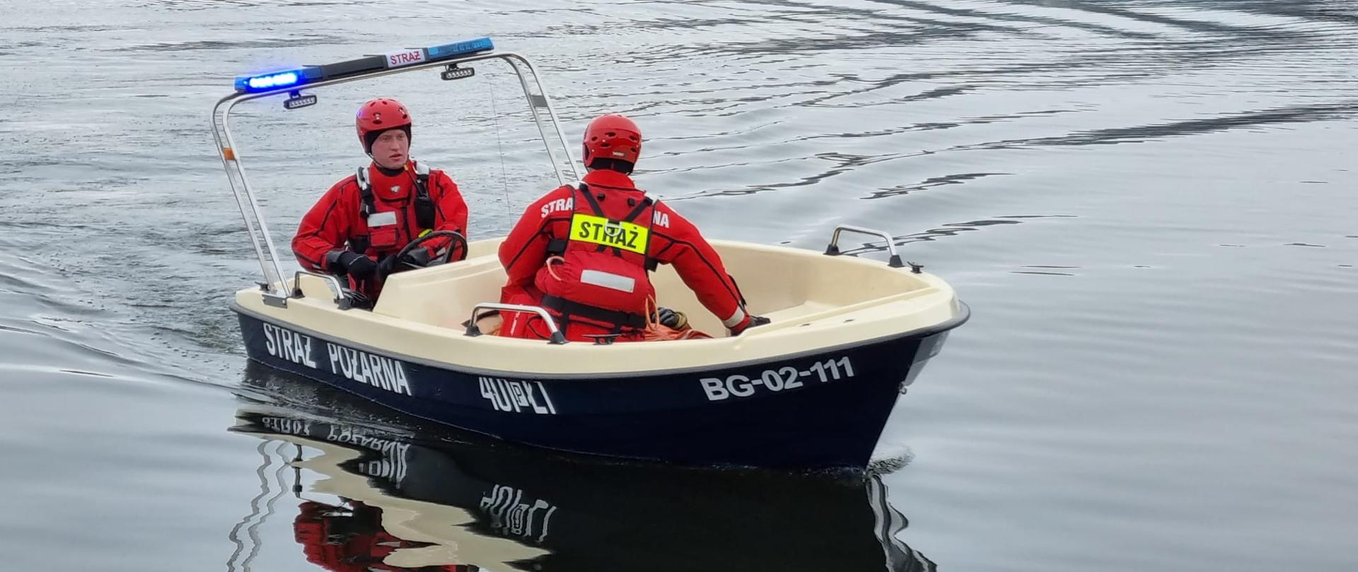 Zdjęcie przedstawia dwóch strażaków ćwiczących na jeziorze ratownictwo wodne – ratowanie poszkodowanego.
W tle jezioro.
