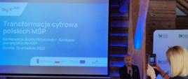 Konferencja pn. „Beskid Przyszłości – fundusze europejskie dla MŚP”, na scenie przemawia do mikrofonu uczestnik wydarzenia, na ekranie napis mówiący o transformacji cyfrowej polskich MŚP
