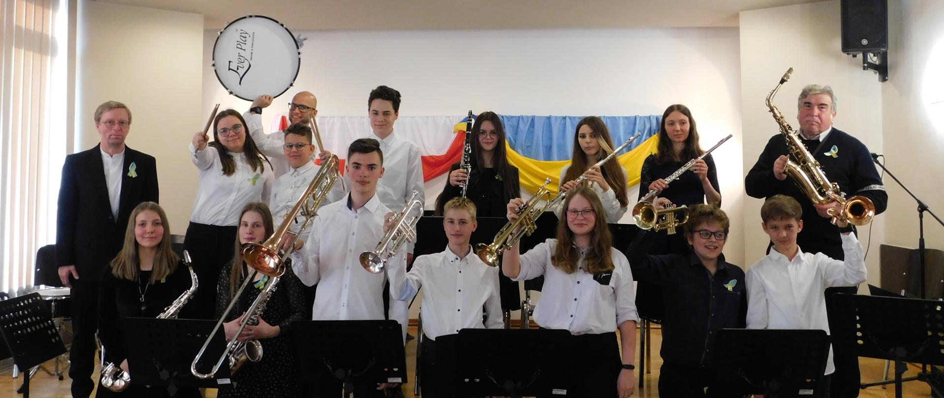 zdjęcie przedstawia uczniów szkoły z instrumentami dętymi: klarnet, saksofon, trąbka, puzon, flet. Po prawej stronie stoi nauczyciel prowadzący orkiestrę, po lewej akompaniator, w oddali widać nauczyciela perkusji z bębnem.