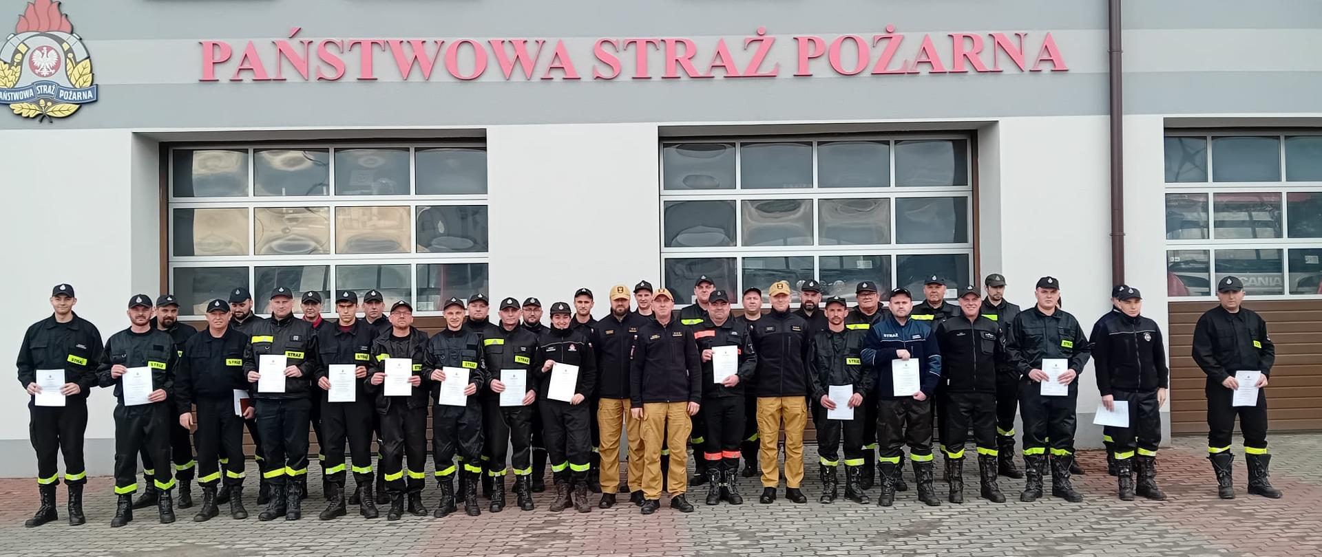 Zdjęcie przedstawia strażaków w ubraniach koszarowych stojących na zbiórce przed budynkiem Jednostki Ratowniczo-Gaśniczej w Lubaczowie.
