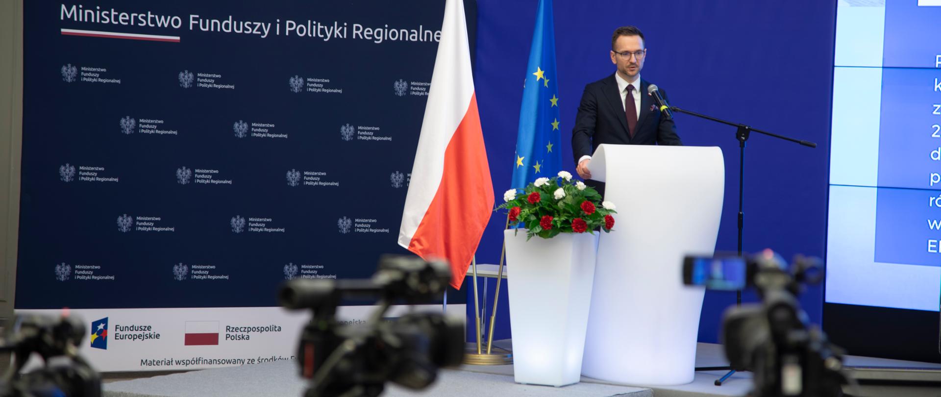 Wiceminister Waldemar Buda stoi w mównicy. Obok niego kwiaty w donicy i flagi UE i PL. Za jego plecami po prawej stronie na ekranie wyświetlona jest prezentacja. Po lewej ścianka z napisem Ministerstwo Funduszy i Polityki Regionalnej. 