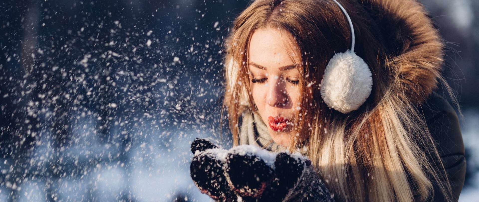 fotografia przedstawia jasnowłosą kobietę ubraną w zimową kurtkę oraz białe nauszniki zdmuchującą z dłoni ubranych w rękawiczki płatki śniegu