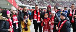 Minister Czarnek w biało-czerwonym szaliku stoi na białej podłodze, wokół niego dzieci w takich samych szalikach, w tle trybuny stadionu.
