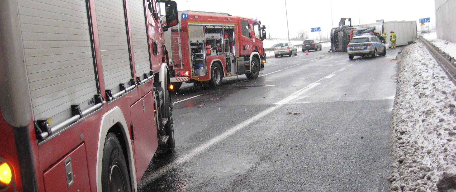 Na zdjęciu znajdują dwa samochody Państwowej Straży Pożarnej, na dalszym planie Policja za nią przewrócona ciężarówka, również na zdjęciu znajdują się dwa samochody osobowe