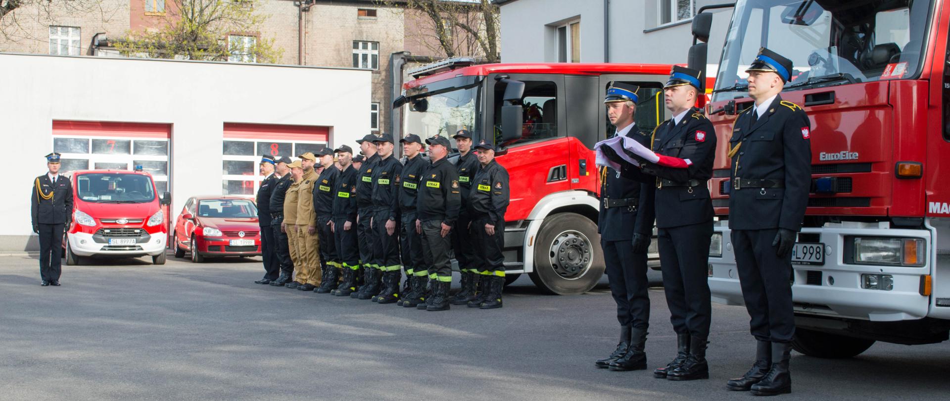 Zdjęcie przedstawia strażaków KM PSP Ruda Śląska podczas uroczystej zmiany służby w związku z obchodzonym w Polsce dniem Flagi Rzeczpospolitej Polskiej. W tle samochody pożarnicze oraz budynek Komendy Miejskiej PSP.