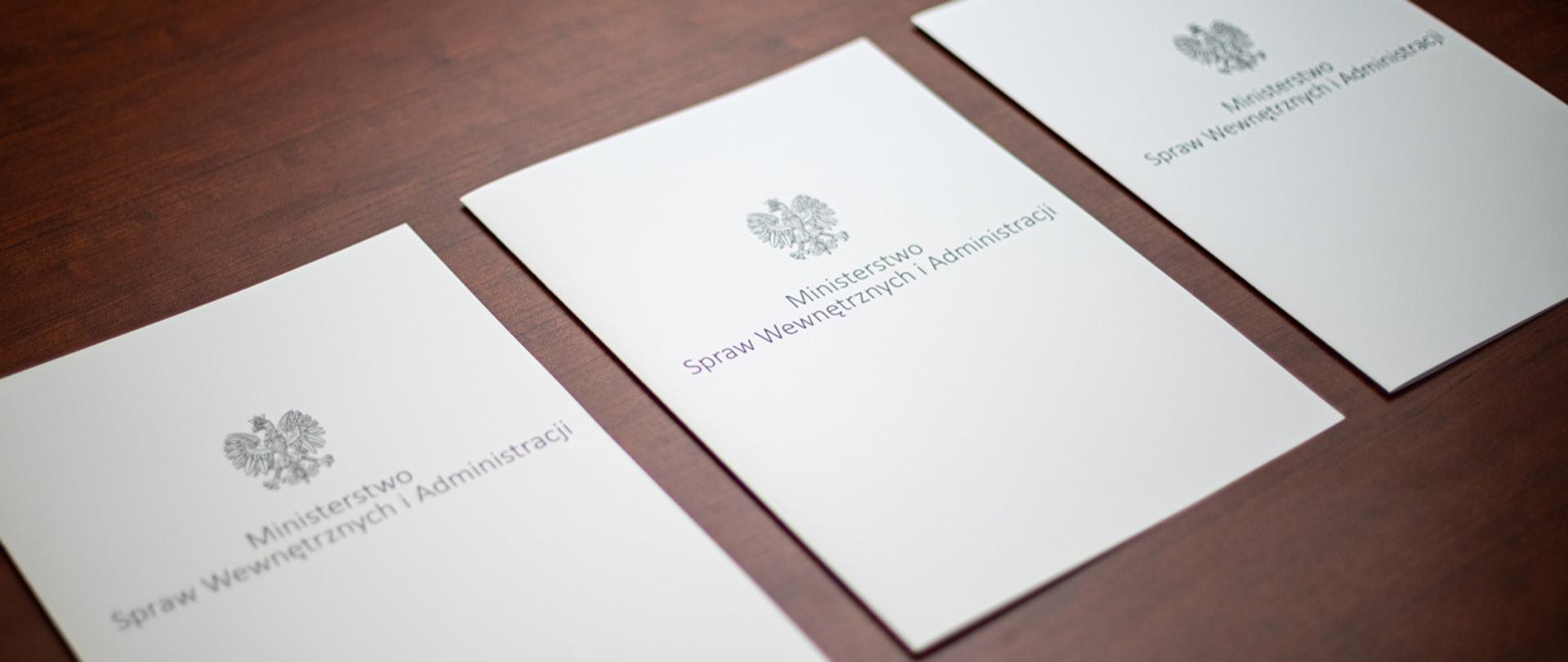 Na zdjęciu widać trzy leżące w rzędzie teczki na dokumenty z napisem "Ministerstwo Spraw Wewnętrznych i Administracji" i orłem z godła narodowego