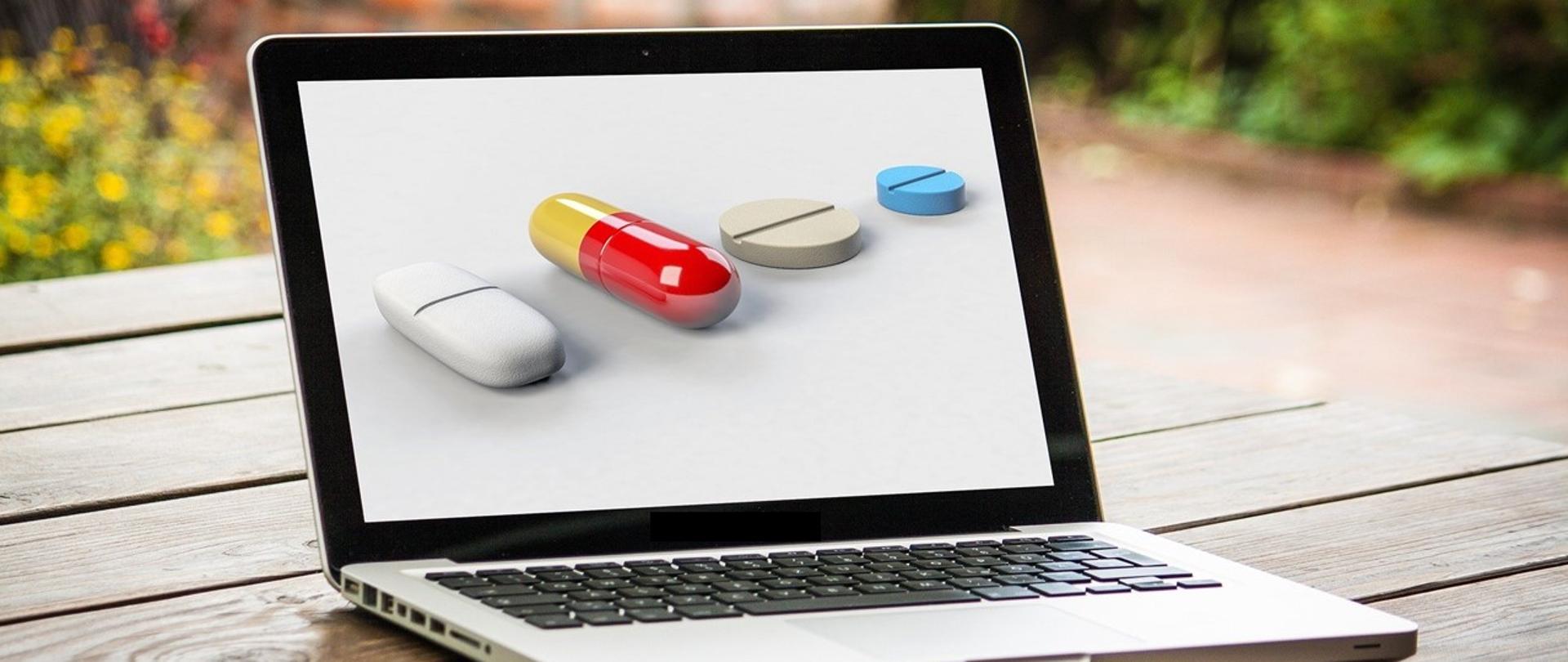 laptop na drewnianym stole, na ekranie różnokolorowe tabletki.