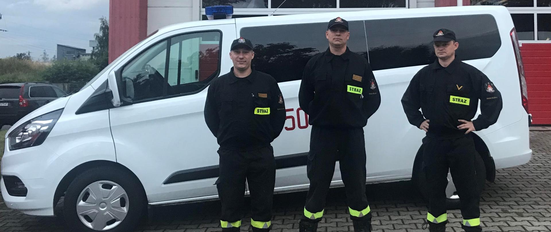trzech strażaków w mundurach koszarowych stoi na tle busa i bramy garażowej