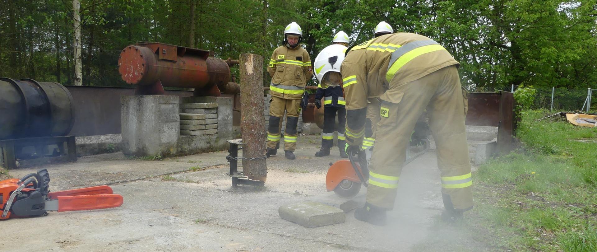 Zdjęcie przedstawia element egzaminu praktycznego kończącego szkolenie podstawowe strażaków ratowników Ochotniczych Straży Pożarnych. Strażacy OSP podczas egzaminu z obsługi pił i pilarek. NA pierwszym planie strażak tnie krawężnik betonowy.