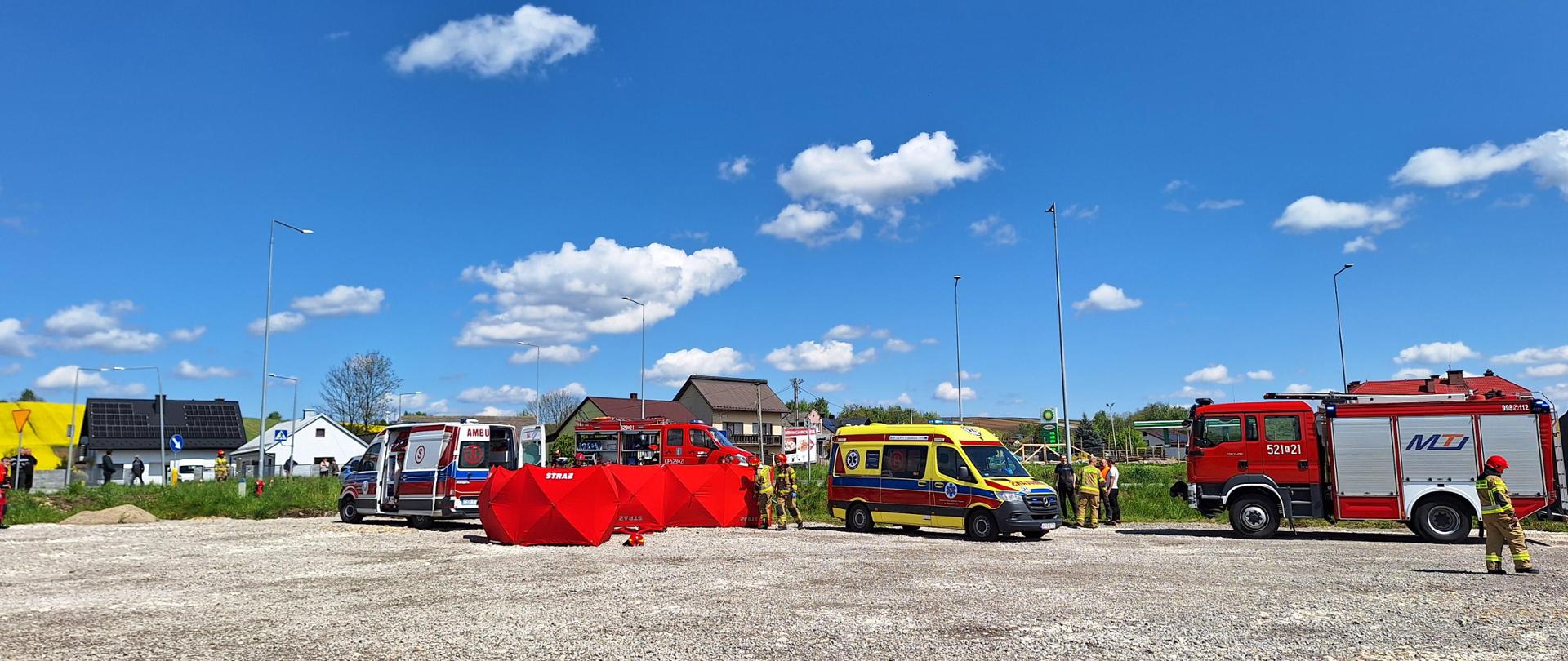 Na zdjęciu stoją samochody strażackie, karetka pogotowia oraz parawany za którymi udzielana jest pierwsza pomoc poszkodowanym
