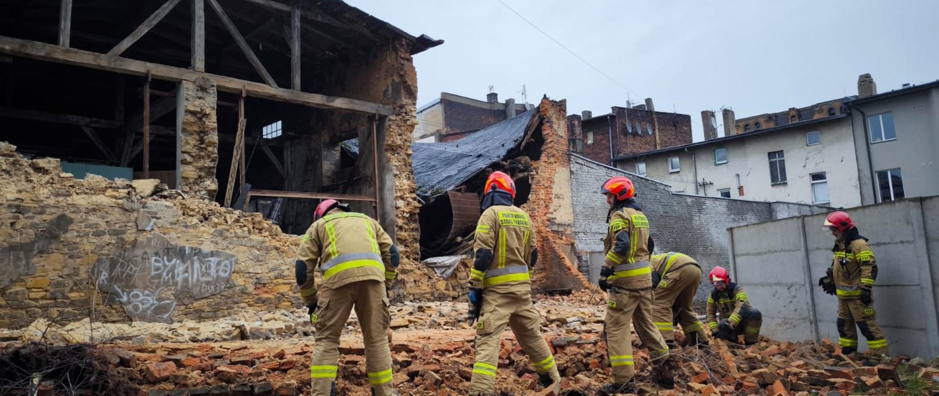 Na pierwszym planie sześciu strażaków przeszukujących gruzowisko, w tle widoczny budynek w którym runęła ściana.