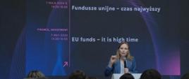 Minister funduszy i polityki regionalnej Katarzyna Pełczyńska-Nałęcz stoi przy pulpicie z mikrofonem, za jej plecami grafika granatowo-fioletowa z napisem "Fundusze unijne – czas najwyższy”
