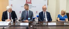 Minister Piontkowski siedzi za stołem, obok kobiety w niebieskiej sukni i dwóch mężczyzn w garniturach.