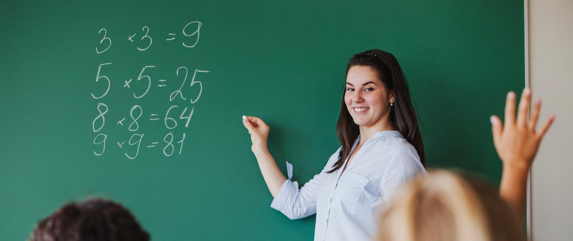 Nauczycielka stoi przy tablicy i pokazuje cyfry 