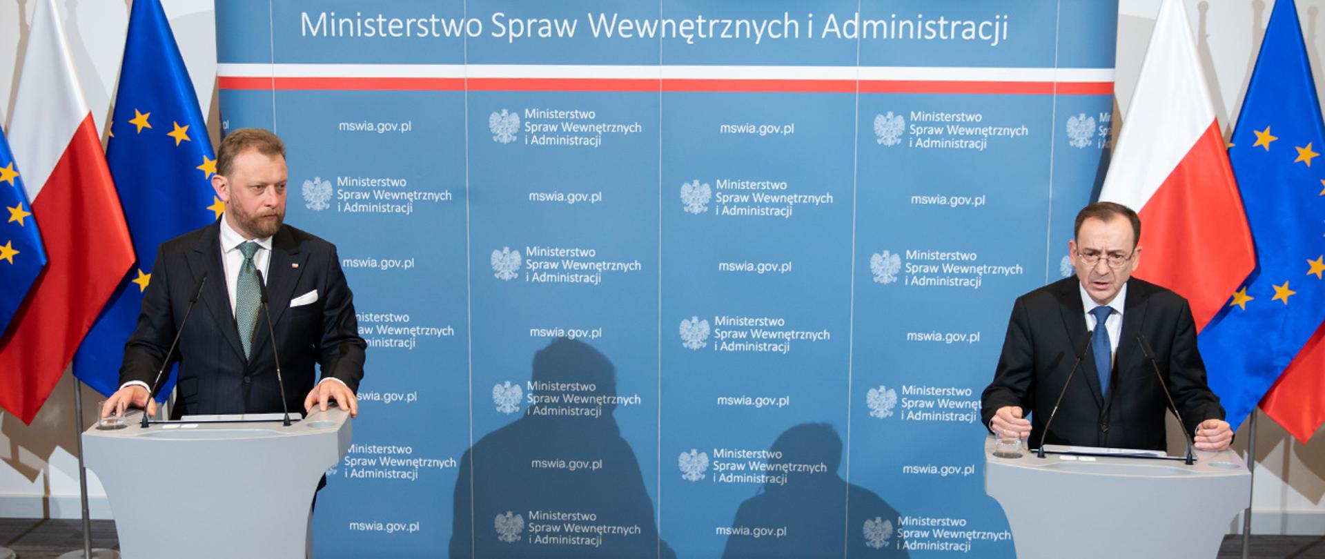 Na zdjęciu widać przemawiającego ministra Mariusza Kamińskiego oraz stojącego obok ministra Łukasza Szumowskiego podczas konferencji prasowej w MSWiA. Obaj stoją na tle ścianki MSWiA i flag Polski i UE.