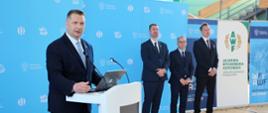 Minister Czarnek stoi przy białej mównicy i mówi do dwóch mikrofonów, za nim stoi trzech mężczyzn w garniturach, za nimi niebieska ściana.