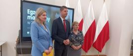 Wiceminister Piontkowski stoi przy kobiecie w jasnoniebieskiej marynarce i kobiecie w bluzce w biało-czarny wzorek, za nimi duży monitor z napisem Egzamin maturalny 2023, dalej na tle białej ściany cztery polskie flagi.