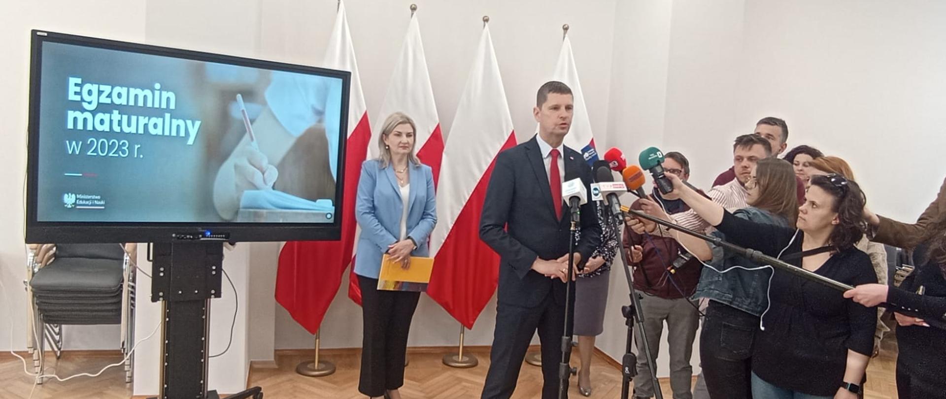 Wiceminister Piontkowski stoi i mówi do mikrofonu na stojaku, przed nim kilkoro dziennikarzy trzymających w rekach mikrofony, za nim monitor z napisem Egzamin maturalny 2023, dalej na tle białej ściany cztery polskie flagi przy których stoi kobieta w jasnoniebieskiej marynarce.