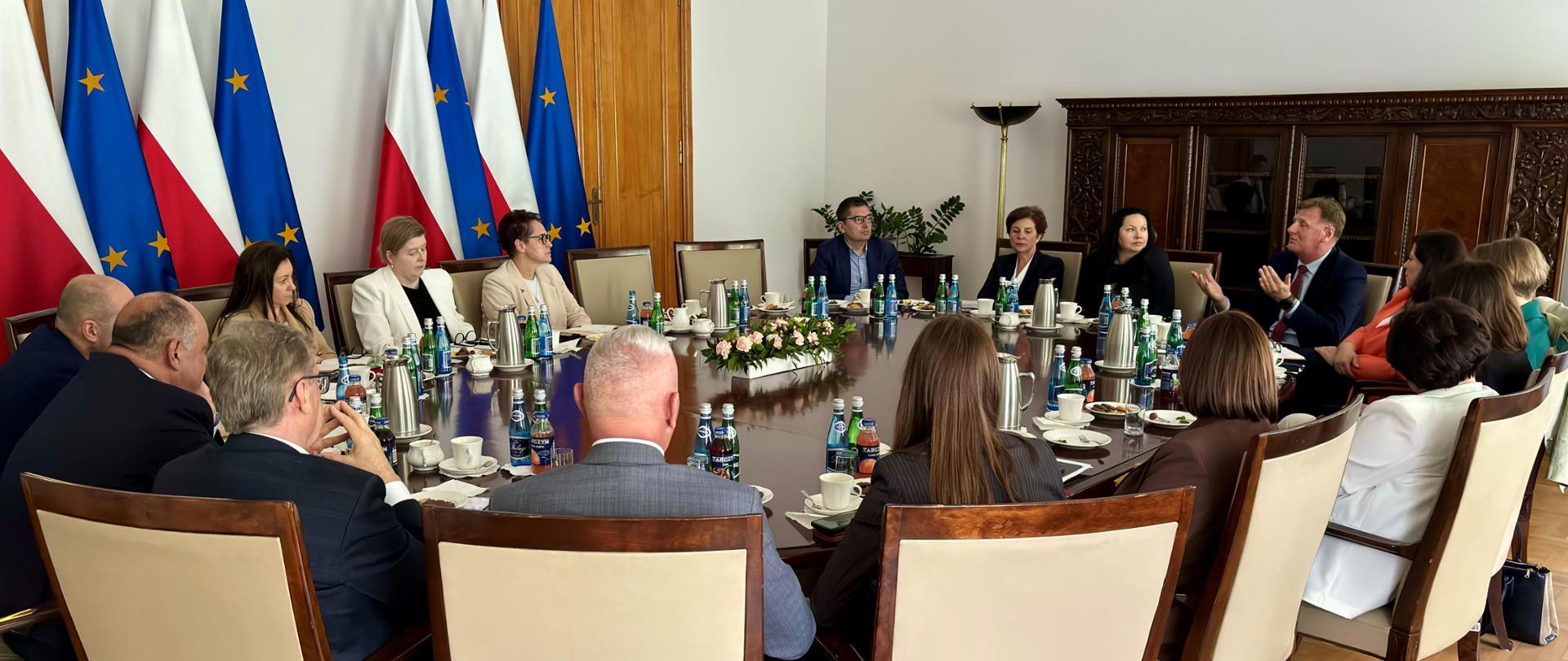 Przy okrągłym stole szefowa służby cywilnej rozmawia z dyrektorami generalnymi urzędów wojewódzkich. Za nimi flagi Polski i Unii Europejskiej.