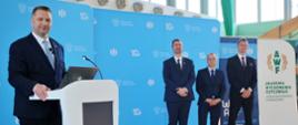 Uśmiechnięty minister Czarnek stoi przy białej mównicy, za nim stoi trzech mężczyzn w garniturach, za nimi niebieska ściana i rozwinięty biały baner z zielonym napisem AWF Akademia Wychowania Fizycznego.