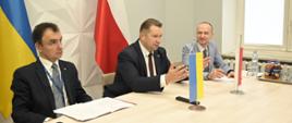 Minister Czarnek i dwóch mężczyzn siedzą przy stoliku, na którym stoją małe flagi Polski i Ukrainy.