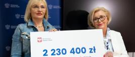 Minister Machałek stoi obok kobiety w niebieskim ubraniu, obie trzymają symboliczny czek z napisem 2 230 400 zł.