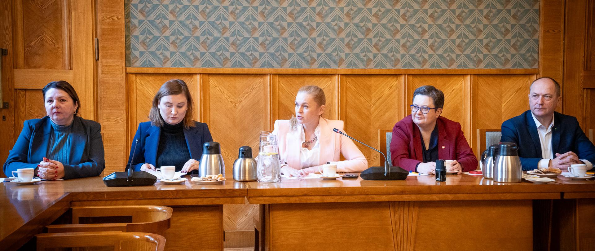 Cztery kobiety i mężczyzna siedzą przy stole. W środku w białej marynarce minister Nowacka mówi do mikrofonu.