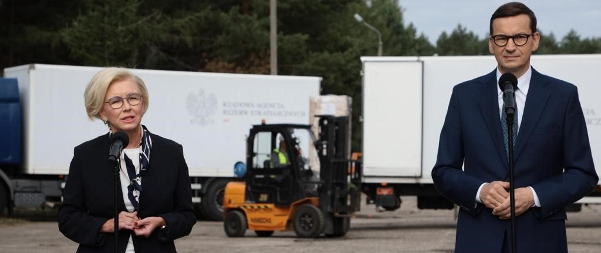 Premier Mateusz Morawiecki i wiceminister Marzena Machałek stoją przed mikrofonami, w tle samochody ciężarowe Rządowej Agencji Rezerw Strategicznych