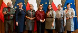 Grupa kobiet i mężczyzn w mundurach harcerskich pozuje do zdjęcia. w tle flagi Polski i UE