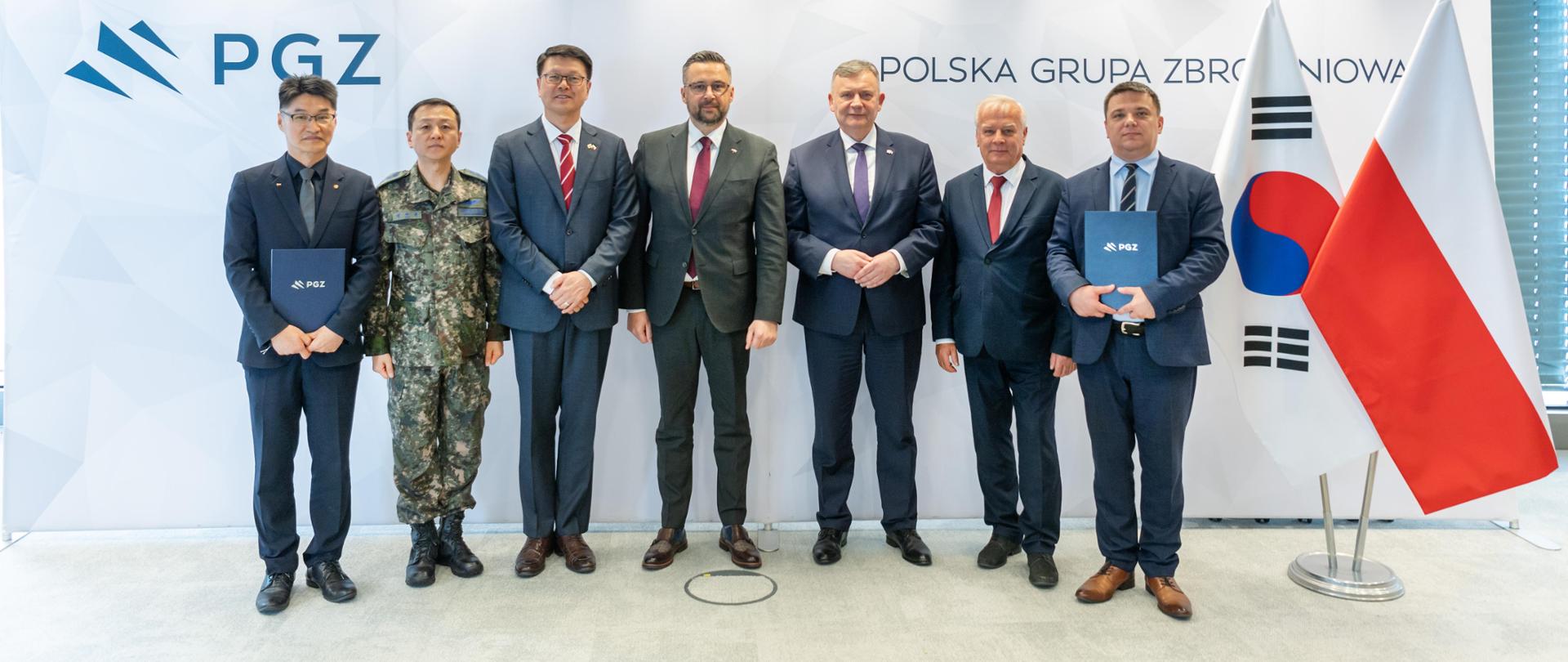 Na zdjęciu stoi siedmiu mężczyzn. W tle ścianka z napisem Polska Grupa Zbrojeniowa i logotyp PGZ. Z prawej strony flagi - Polski i Korei Południowej.