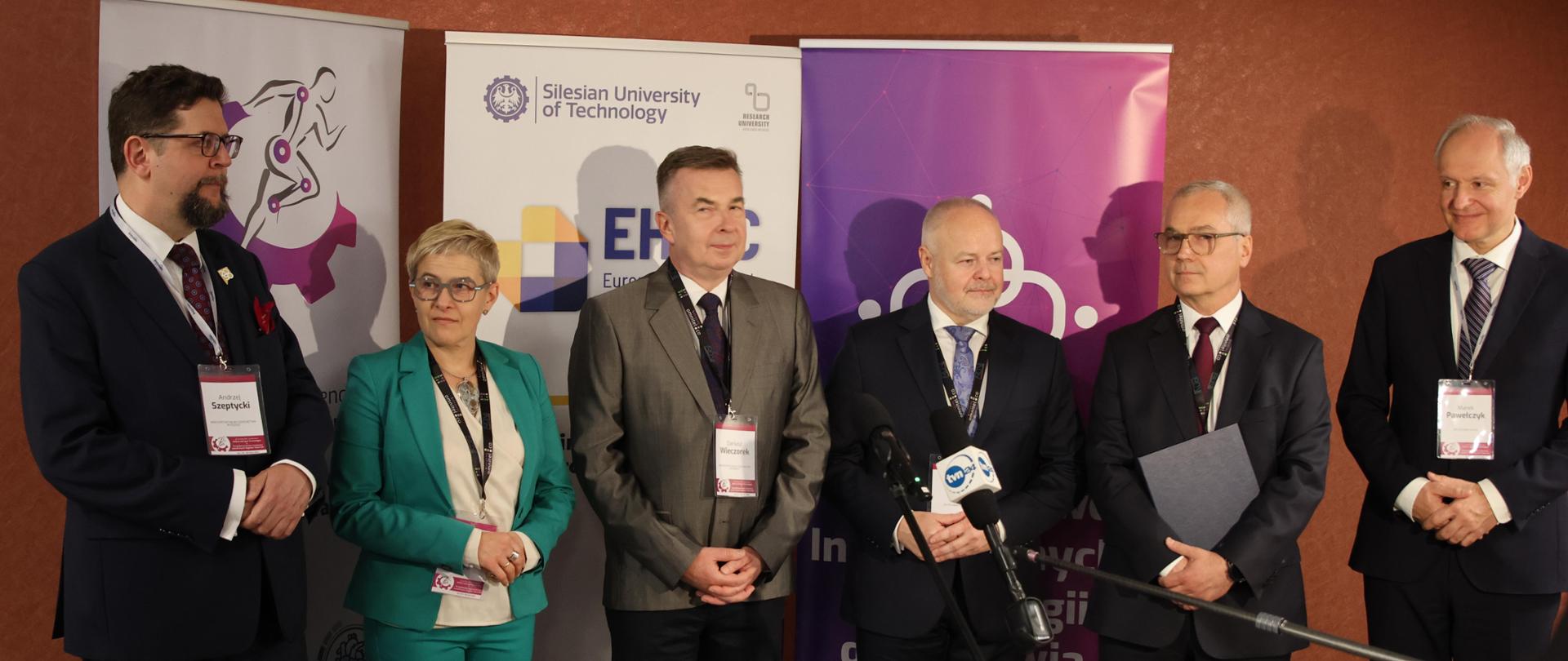 Minister Dariusz Wieczorek z Wiceministrami: Marią Mrówczyńską, Andrzejem Szeptyckim, Markiem Gzikiem oraz dwoma mężczyznami stoją przed mikrofonem.