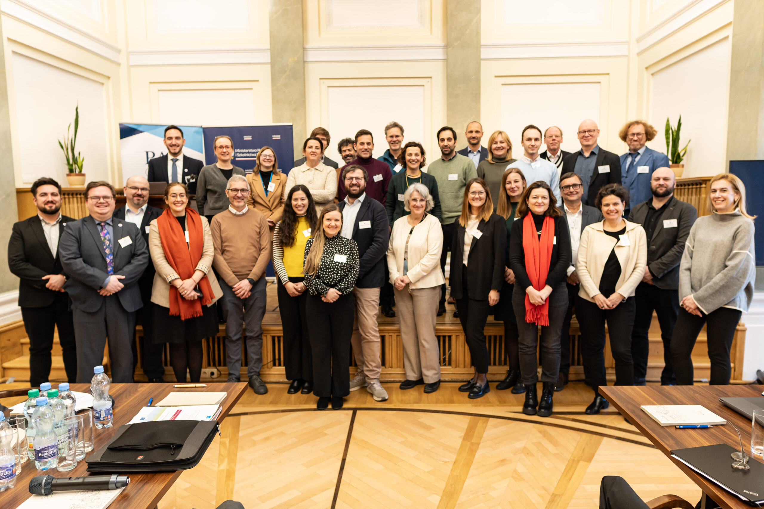 Zdjęcie zbiorowe uczestników spotkania - na tle żółtej ściany na podłodze z drewnianych klepek stoi grupa kilkudziesięciu kolorowo uubranych osób.