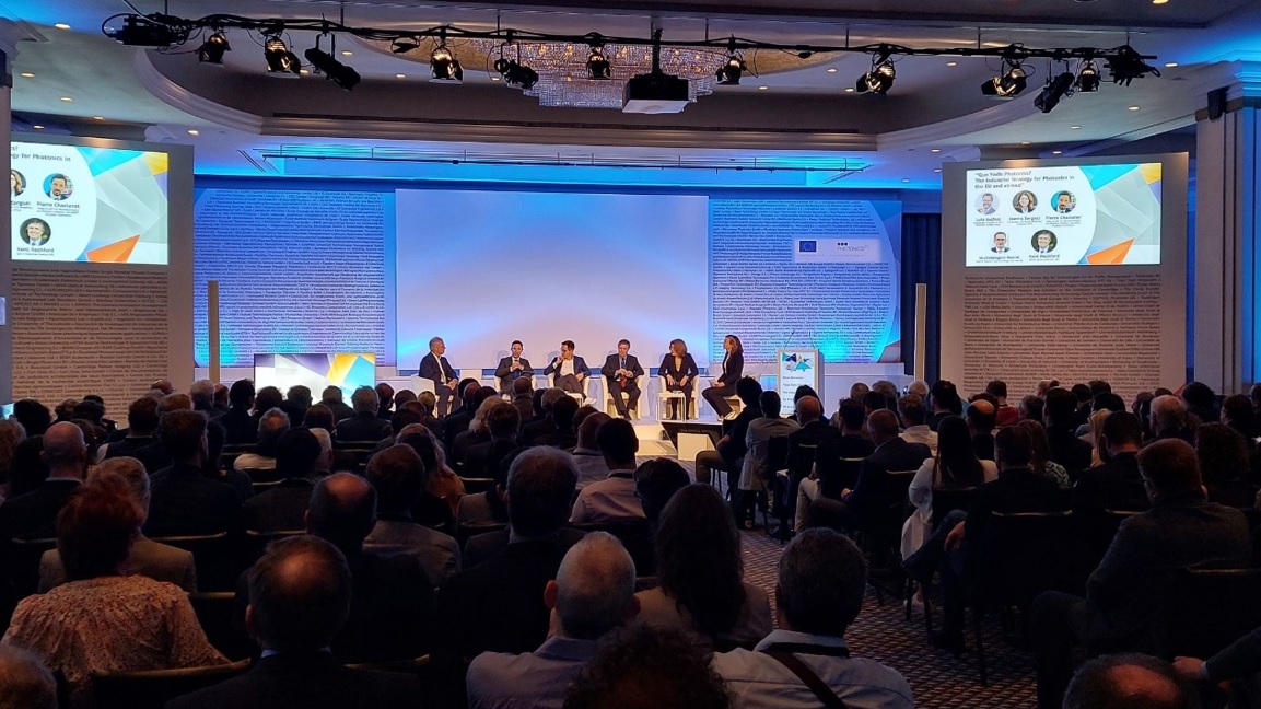 Na zaciemnionej sali w rzędach krzeseł siedzą ludzie, przed nimi pod ścianą na fotelach siedzi kilka rozmawiających osób.