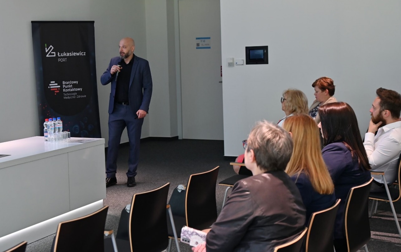 Na sali w rzędach krzeseł siedzą ludzie, przed nimi mężczyzna w garniturze mówi do mikrofonu.