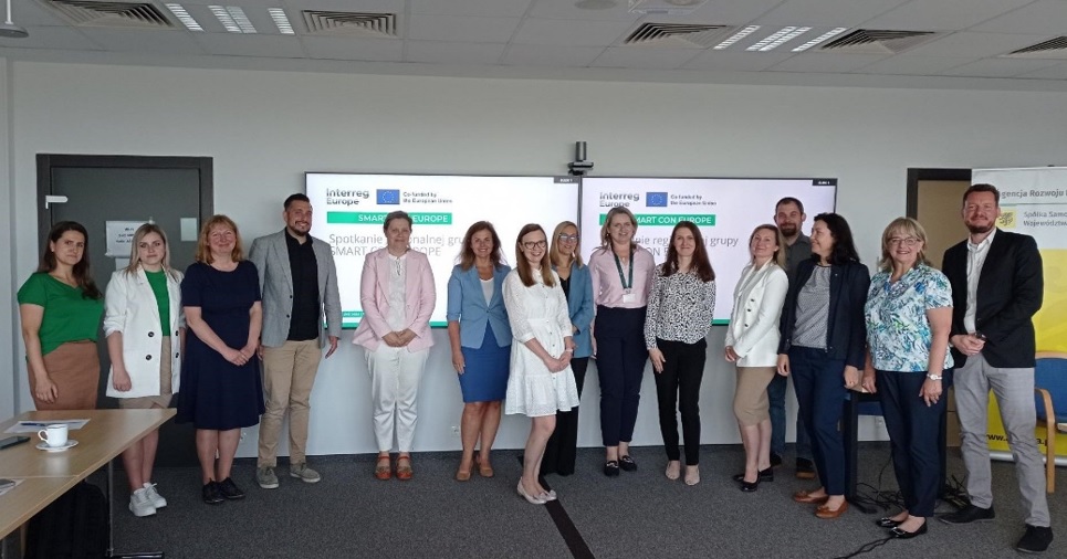 Zdjęcie zbiorowe, na sali pod ścianą na której wiszą dwa duże monitory stoi w szeregu 15 osób.