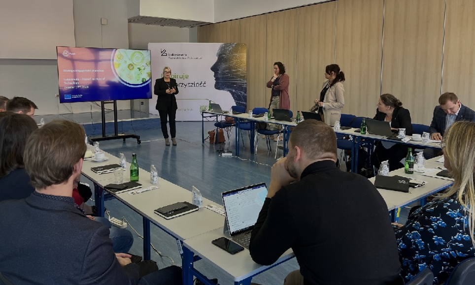 Na sali przy ustawionych w czworokąt stołach siedza ludzie, przed nimi kobieta w czarnej marynarce stoi i mówi do mikrofonu.