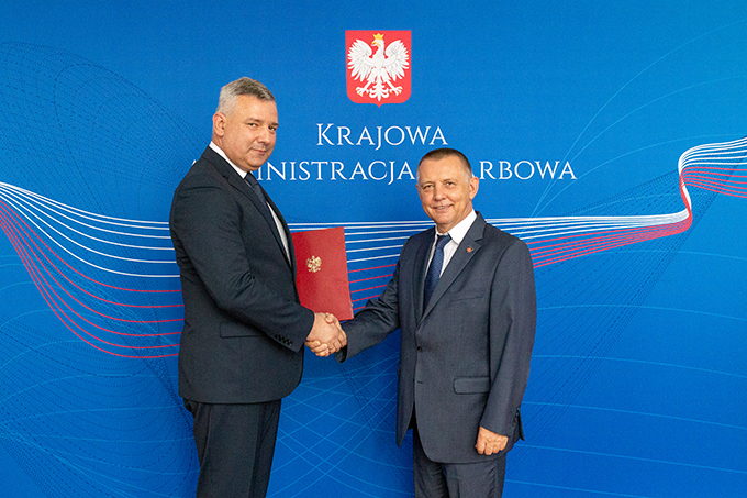 Uśmiechnięci minister finansów Marian Banaś i minister Piotr Dziedzic podają sobie ręce na tle banera Krajowej Administracji Skarbowej