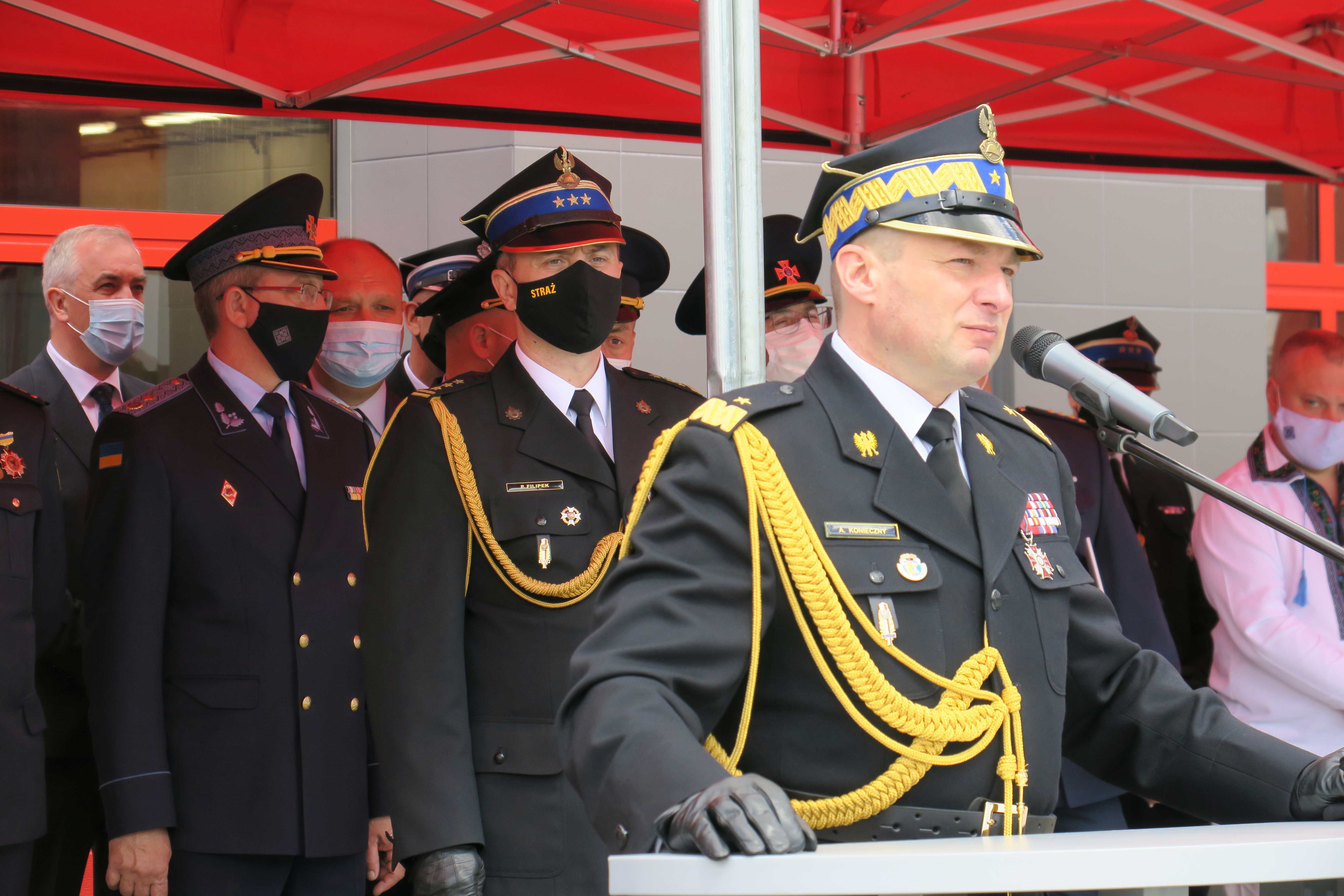 Trybuna honorowa na której pod czerwonym baldachimem stoją strażacy w mundurach galowych. Przemawia zastępca komendanta głównego PSP