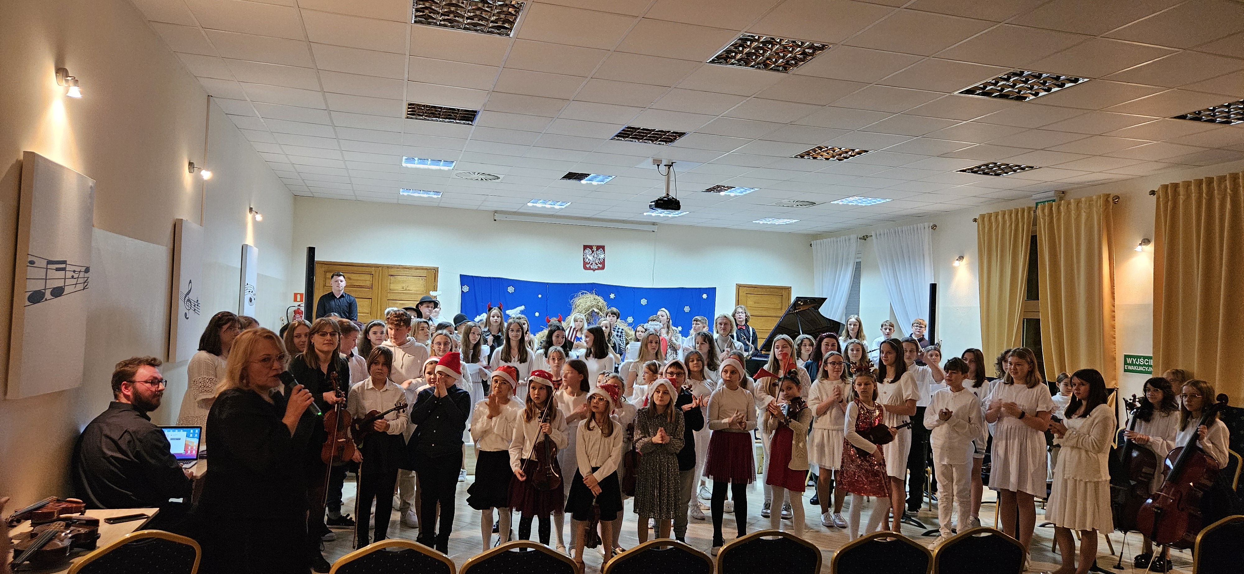 Zdjęcie przedstawia wszystkich wykonawców, którzy wzięli udział w jasełkach, stojących przed sceną i na scenie. Po lewej stronie przemawia pani Dyrektor Agata Świderska. W tle scenografia imitująca niebo. Po bokach z prawej strony-zasłony w kolorze żółtym oraz z lewej grafiki biało czarne.