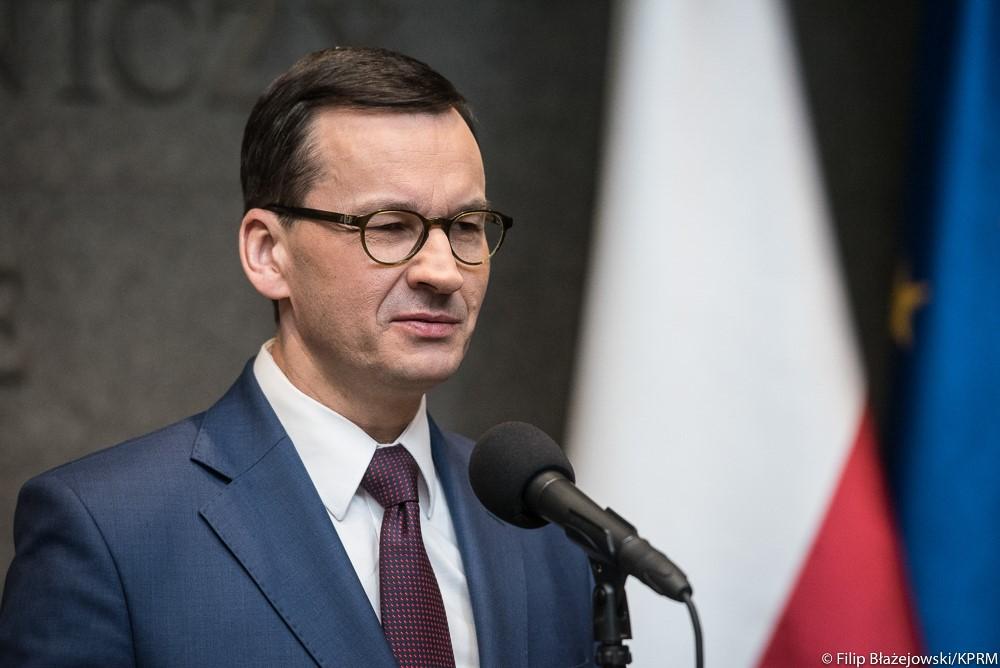 Premier Mateusz Morawiecki przy mikrofonie, a w tle flagi Polski i UE.