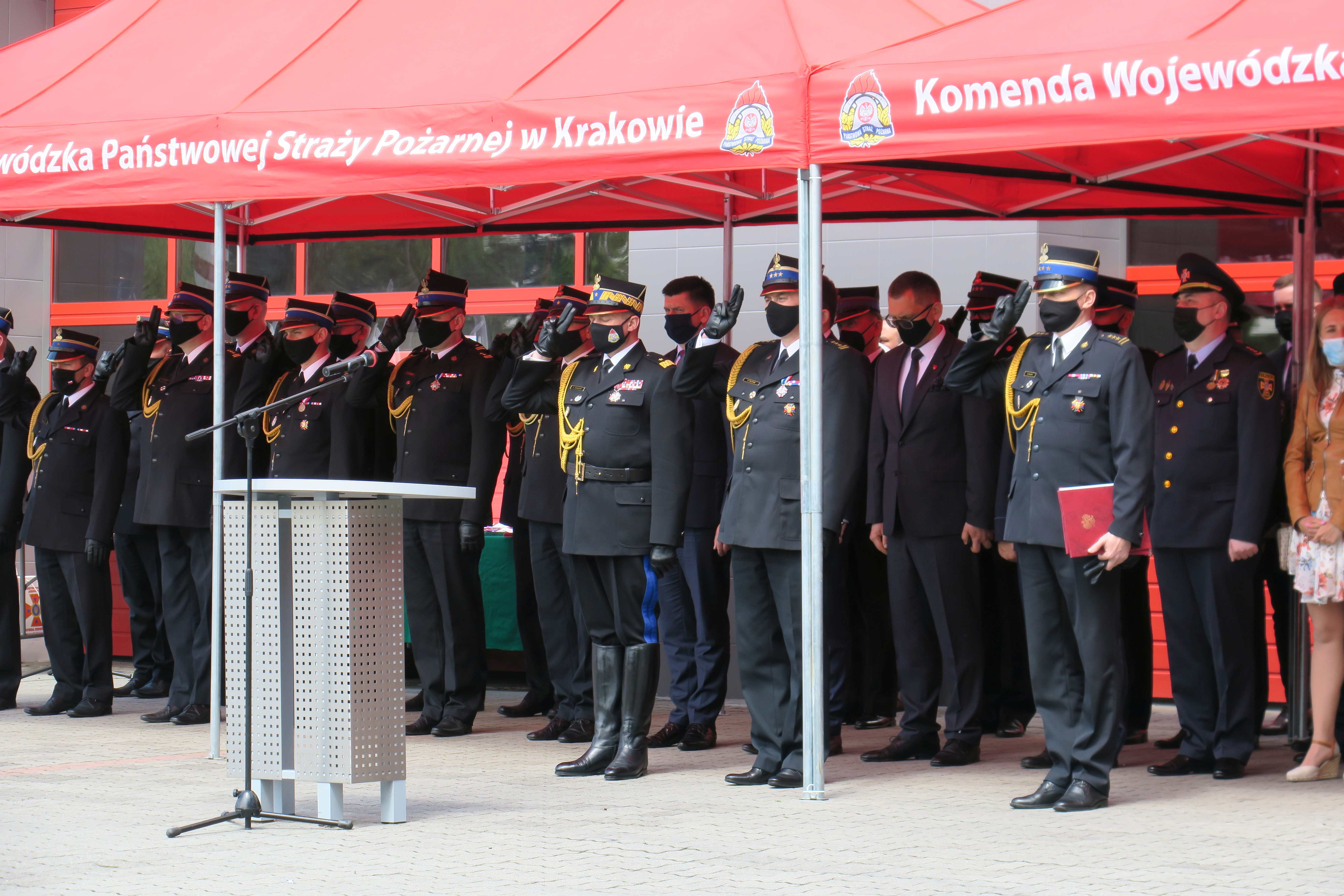Trybuna honorowa na której pod czerwonym baldachimem stoją strażacy w mundurach galowych i oddają honor.