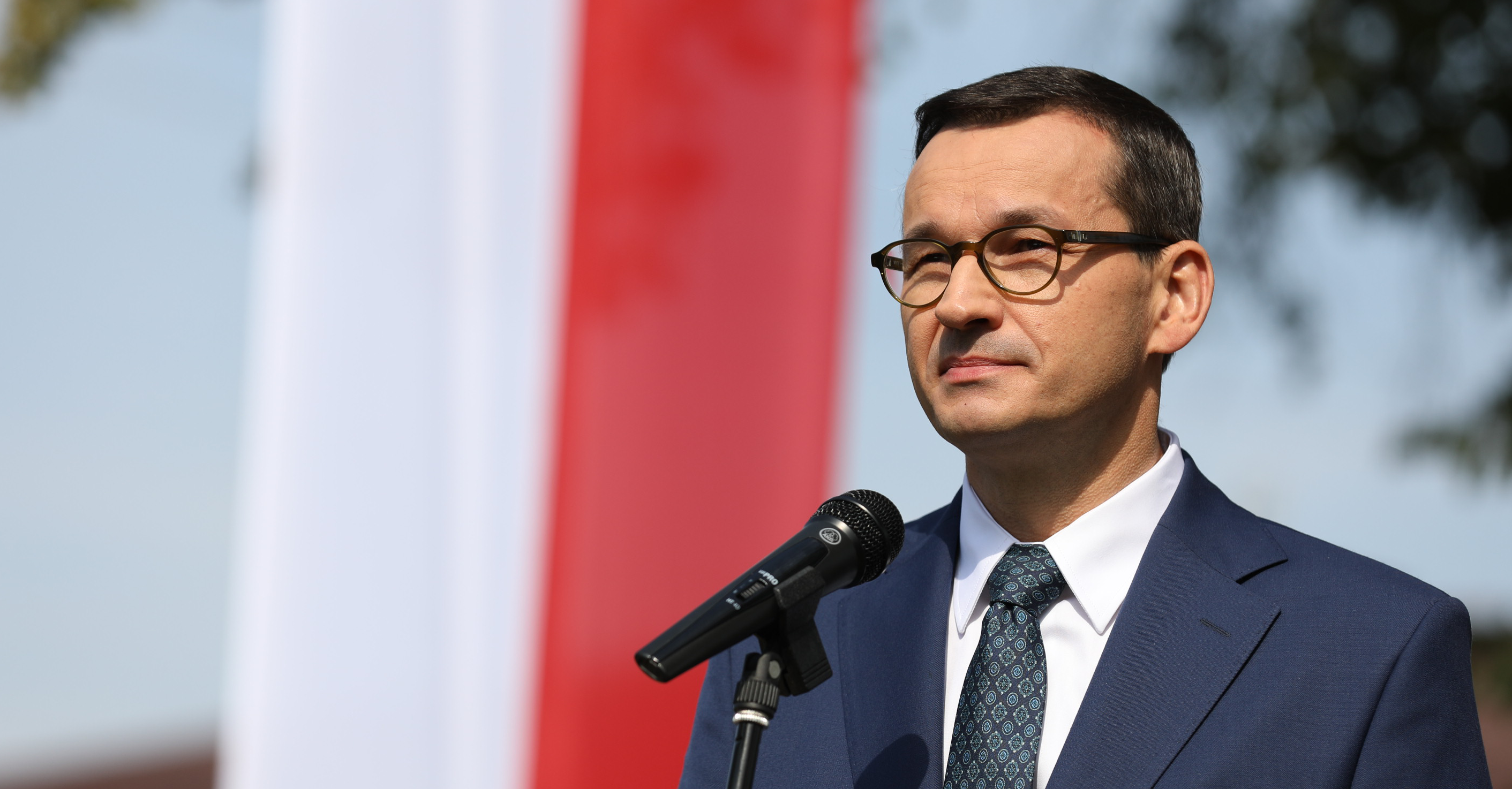 Premier Mateusz Morawiecki podczas przemówienia na tle flagi.