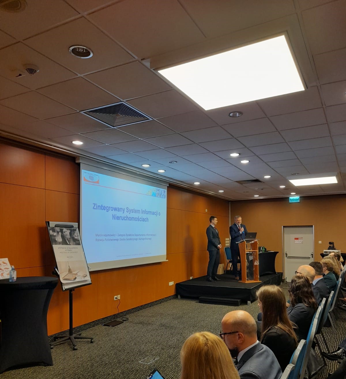 Zdjęcie przedstawia Głównego Geodetę Kraju dr. hab. inż. Waldemara Izdebski oraz Zastępcę Dyrektora Departamentu Departamentu Informatyzacji i Rozwoju Państwowego Zasobu Geodezyjnego i Kartograficznego - Marcina Adamowicza prezentującego informacje na temat ZSIN.